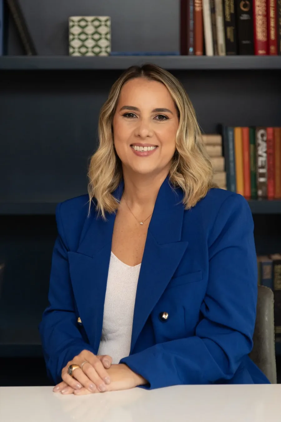 Cristiane Barreto, business and career consultant, seated at a white desk in a royal blue blazer, with her office library of business books behind her